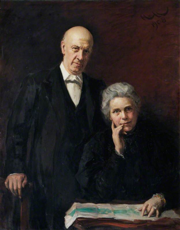 An older, bald man in a suit stands beside a seated older woman with gray hair, who rests her hand on her chin. Both face forward, with a map or paper on the table in front of them, against a dark background.
