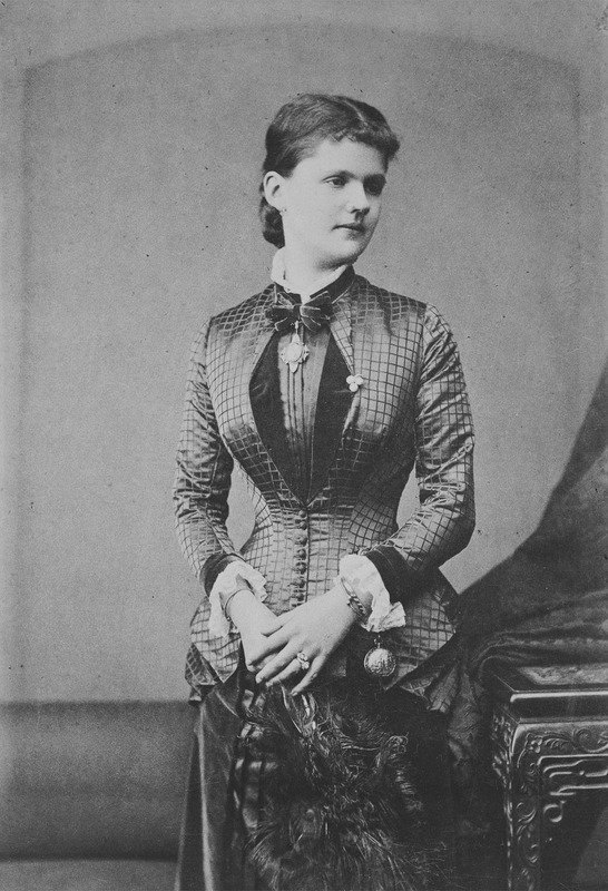 Black-and-white photo of a young woman in a patterned, high-necked Victorian dress, standing beside an ornate table, holding dark feathers, and looking slightly to her left.