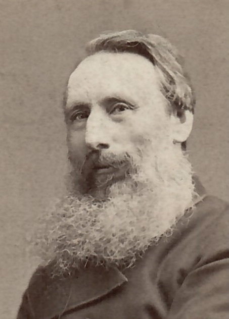 A sepia-toned portrait of an older man with a full, bushy white beard and mustache, wearing a dark coat, looking slightly to the side against a plain background.