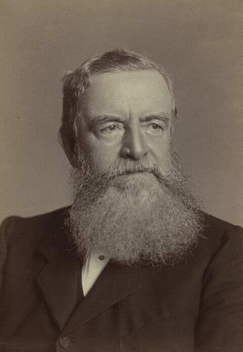 Portrait of an older man with a large, full white beard, short hair, and a serious expression. He is wearing a dark suit jacket and looking slightly to the side against a plain background.