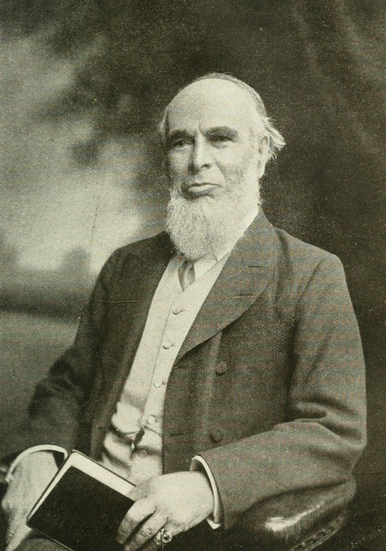 An older man with a white beard sits in a chair, wearing a suit and holding a closed book in his lap. The photo is black and white with a blurred background.